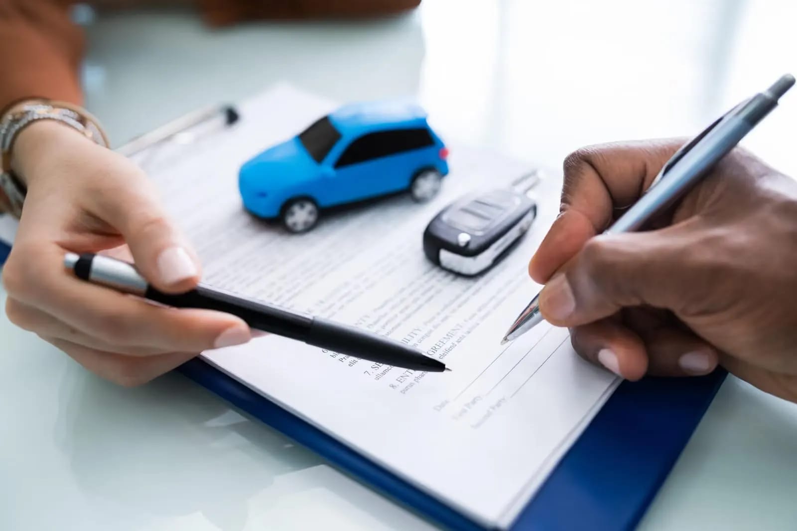 Duas pessoas assinando um contrato de seguro de carro, com uma miniatura de carro azul e uma chave sobre a mesa